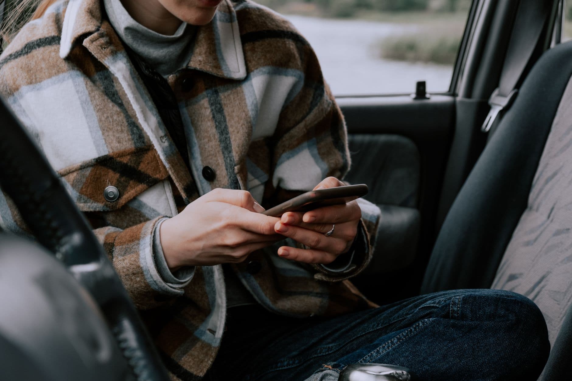 An individual seated in a vehicle using a smartphone, wearing a checkered jacket.